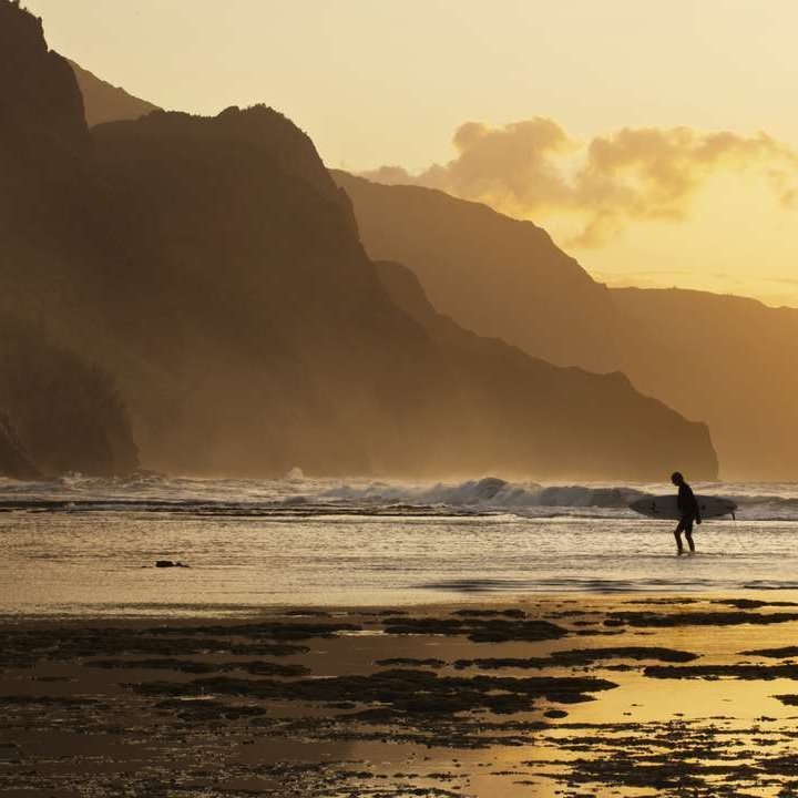 Un surfeur marche sur la plage à Hawaï, avec un magnifique coucher de soleil en arrière-plan.