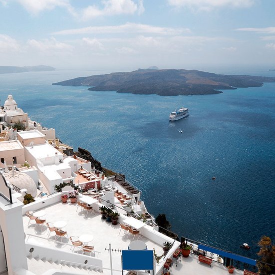 Mittelmeerblick mit weissen Terrassen und Kreuzfahrtschiff vor griechischer Inselkulisse.