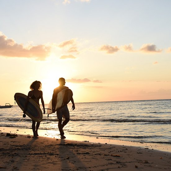 Surfer laufen bei Sonnenuntergang am Strand entlang – sportliche Aktivferien weltweit mit Kuoni Sports.