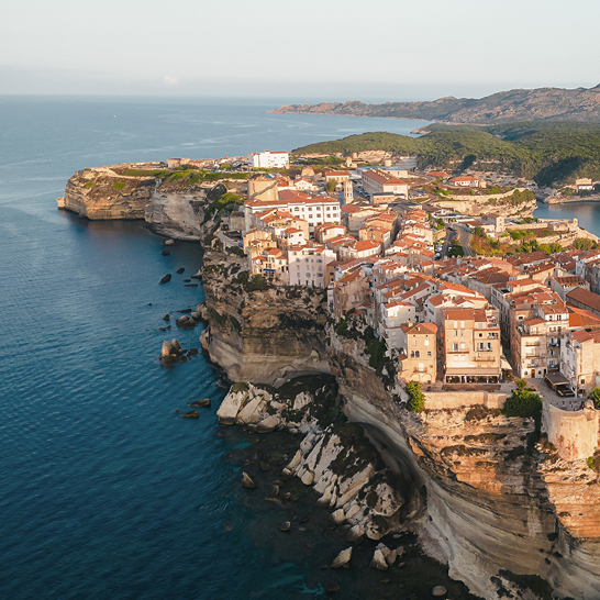 Vue aérienne d'une ville perchée sur une falaise surplombant l'océan, avec des maisons colorées et des vagues en dessous.