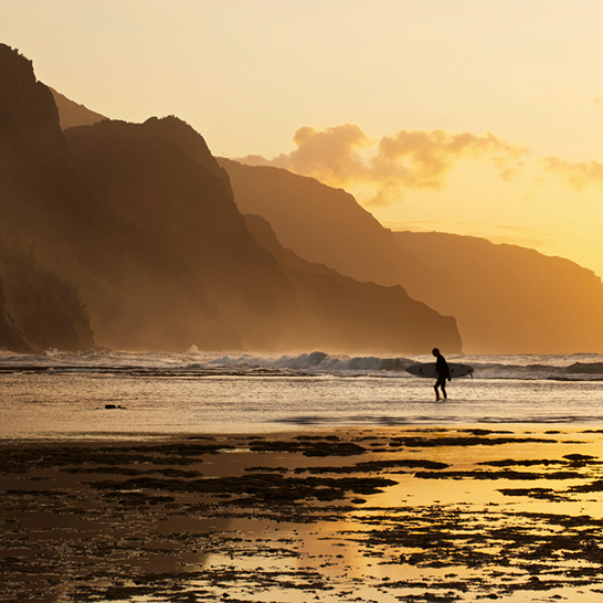 Une personne marche sur la plage à Hawaii, portant une planche de surf sous le bras.